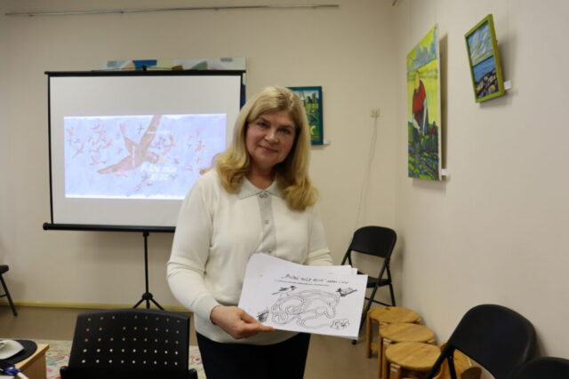 RCB filiālbibliotēkas "Strazds" un Grīziņkalna filiālbibliotēkas vadītāja Jeļena Kuzņecova rāda pasākuma apmeklētājiem sagatavotos mājasdarbus